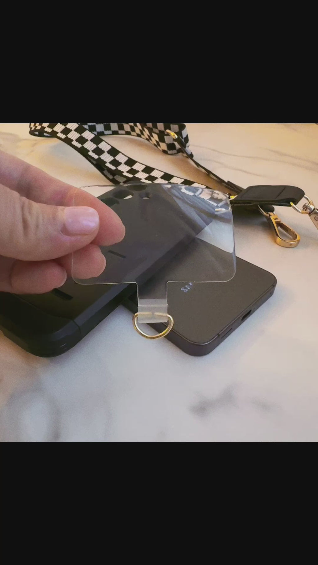 Person holding a clear phone lanyard attachment tab over a black phone on a marble surface.