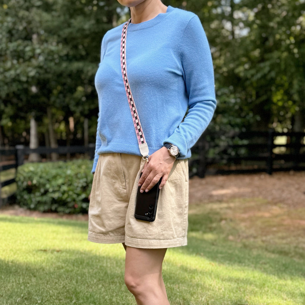 Person wearing a blue sweater and beige shorts standing outdoors with greenery in the background  using checkered crossbody phone strap