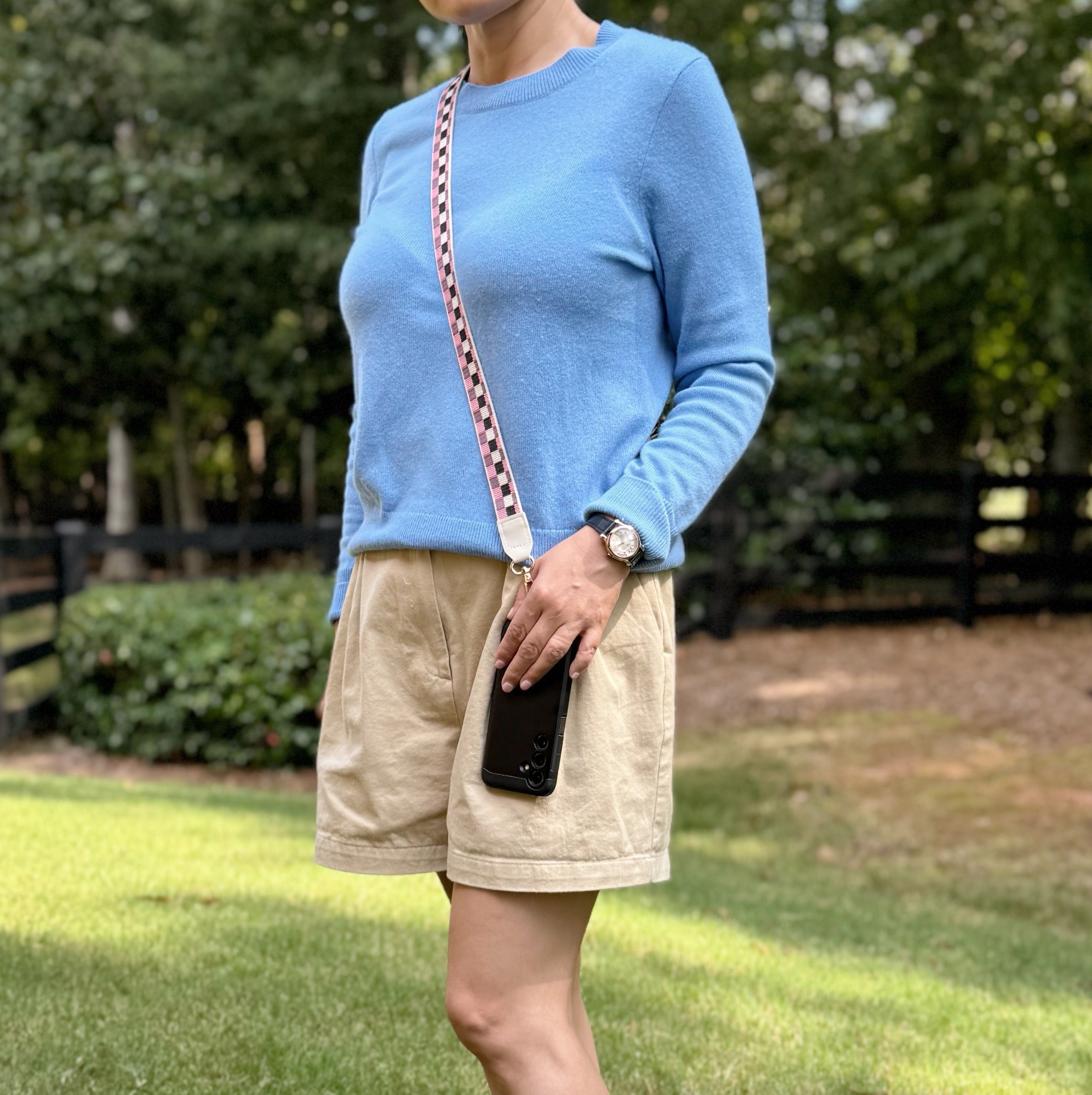 Person wearing a blue sweater and beige shorts standing outdoors with greenery in the background  using checkered crossbody phone strap